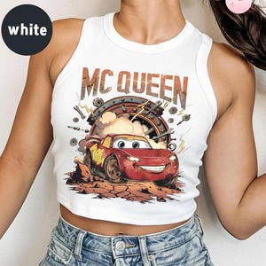 May include: White crop top featuring a graphic of Lightning McQueen from the film Cars. The design includes the text "MC QUEEN" above the car, set against a backdrop of tools and a circular frame.