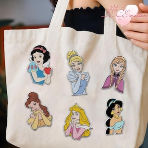 May include: A white tote bag with six colorful embroidered patches of Disney princesses. The princesses include Snow White, Cinderella, Anna, Belle, Aurora, and Jasmine. Each patch features a detailed illustration of the character.