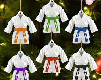May include: White karate uniform ornaments with colored belts: orange, green, blue, purple, red, and black. Each ornament has "2025" and "HENRY" printed on the belt and shorts. Gold string for hanging. Set against a green Christmas tree.