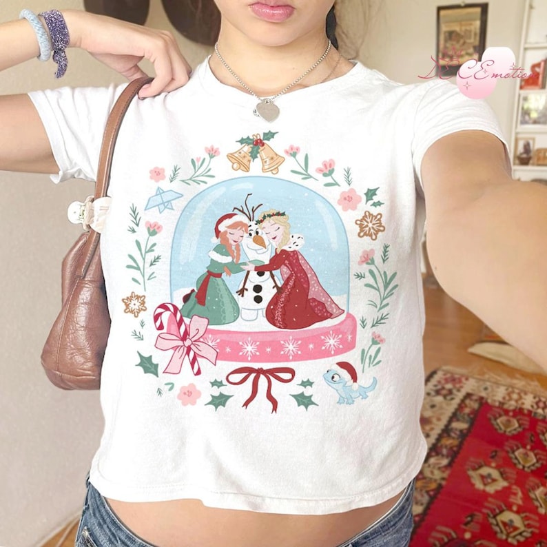 May include: White t-shirt featuring a snow globe design with the characters Anna, Elsa, and Olaf from the movie Frozen. The design includes floral and snowflake accents, a candy cane, and a small blue dragon.