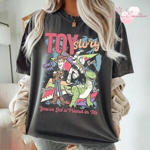 May include: Dark gray t-shirt featuring a colorful graphic from the movie "Toy Story." The design includes Woody, Buzz Lightyear, and Rex, with the text "TOY STORY" and "You've Got a Friend in Me." The shirt also has "SINCE 1995" printed on it.