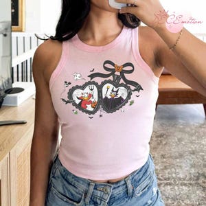 Puede incluir: Camiseta sin mangas rosa claro con ribete rosa claro. Presenta un dise&ntilde;o de Halloween con dos marcos en forma de coraz&oacute;n, personajes de dibujos animados, murci&eacute;lagos y un lazo negro con una calabaza.