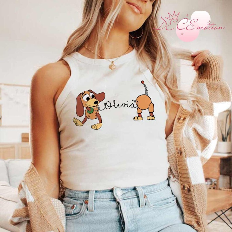 May include: A white tank top featuring a cartoon illustration of a dachshund dog with the name "Olivia" in cursive. The dog is brown and tan with a green collar. The tank top is sleeveless.