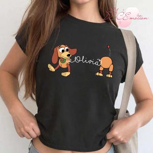 May include: Black t-shirt featuring a cartoon illustration of a dachshund dog with the name "Olivia" in cursive script. The dog is depicted in two parts, with the front and back halves separated.
