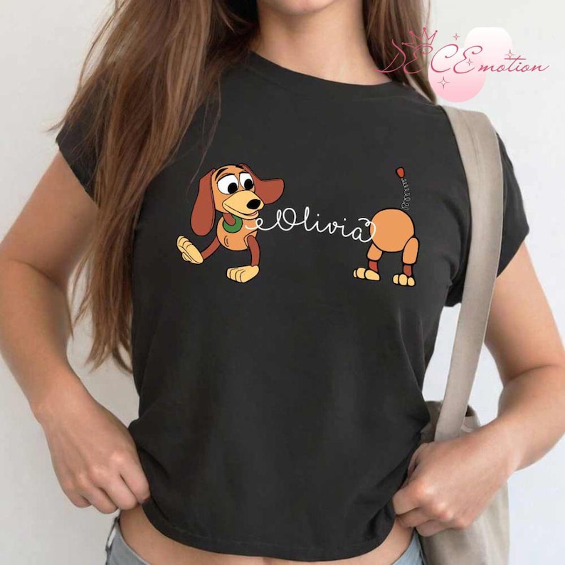 May include: Black t-shirt featuring a cartoon illustration of a dachshund dog with the name "Olivia" in cursive script. The dog is depicted in two parts, with the front and back halves separated.