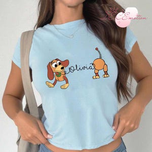 May include: Light blue t-shirt featuring a cartoon dog design. The design includes a brown and orange dog with the name "Olivia" in cursive. The dog is in a playful pose, with its tail raised.