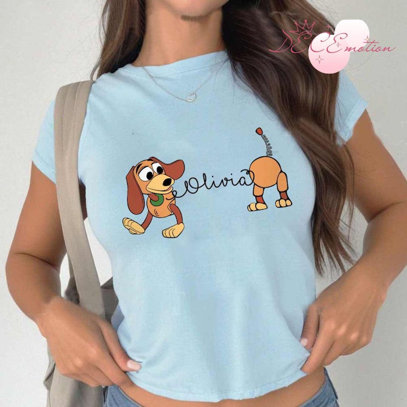 May include: Light blue t-shirt featuring a cartoon dog design. The design includes a brown and orange dog with the name "Olivia" in cursive. The dog is in a playful pose, with its tail raised.