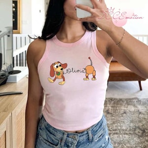 May include: Pink tank top with a light pink trim featuring a cartoon dog graphic and the name "Olivia". The dog is brown, tan, and yellow. The tank top is made of a soft material.