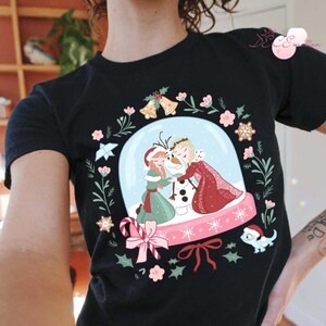 May include: Black t-shirt featuring a snow globe design with the characters Anna, Elsa, and Olaf from the movie Frozen. The snow globe is surrounded by a wreath of flowers, bells, and snowflakes. A small blue lizard wearing a Santa hat is also included in the design.