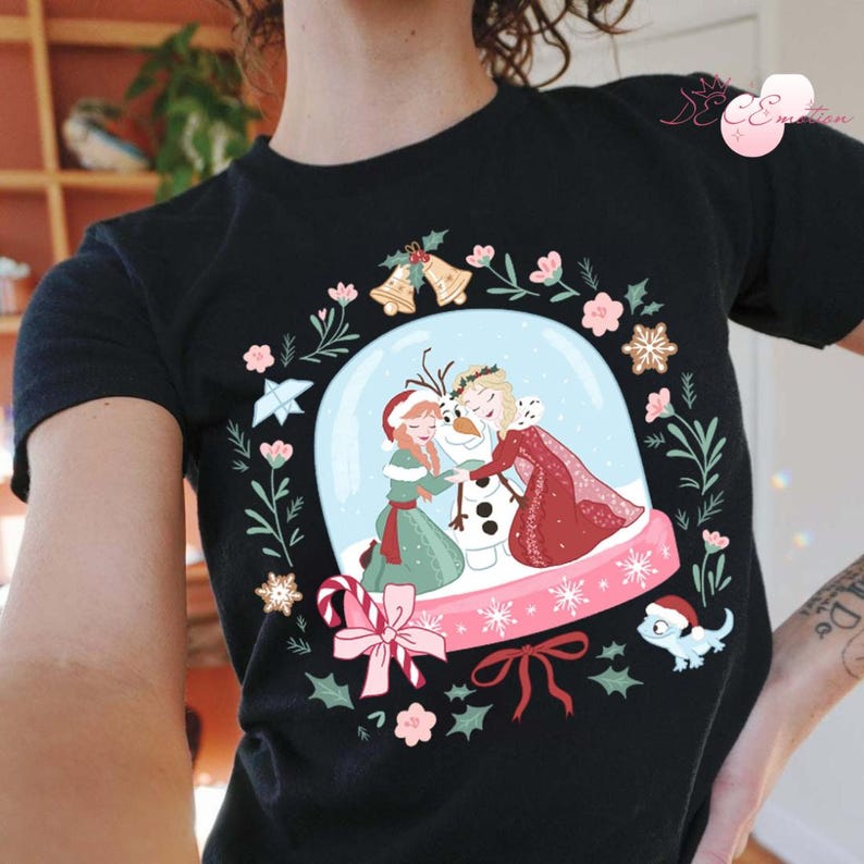 May include: Black t-shirt featuring a snow globe design with the characters Anna, Elsa, and Olaf from the movie Frozen. The snow globe is surrounded by a wreath of flowers, bells, and snowflakes. A small blue lizard wearing a Santa hat is also included in the design.