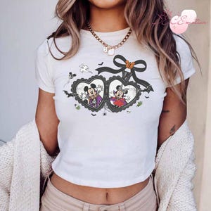 Puede incluir: Camiseta blanca con un dise&ntilde;o de Halloween. El dise&ntilde;o presenta a Mickey y Minnie Mouse disfrazados dentro de marcos en forma de coraz&oacute;n, un lazo negro con una calabaza, murci&eacute;lagos y fantasmas.