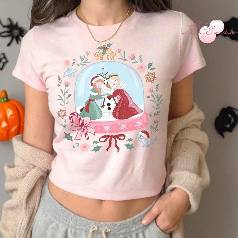 May include: A light pink t-shirt featuring a snow globe design with cartoon characters. The snow globe contains two figures hugging a snowman. The design is surrounded by floral and holiday-themed elements.