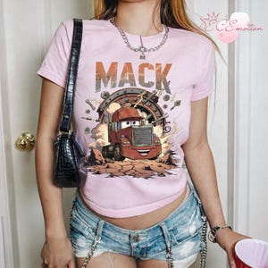 May include: A pale pink t-shirt with a cartoon image of the character Mack from the film Cars. The word "MACK" is printed above the truck. The design includes a red truck, gears, and lightning bolts.