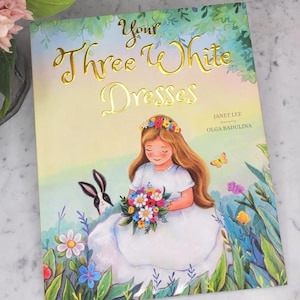 May include: A children's book cover titled "Your Three White Dresses" with gold lettering. The illustration features a girl in a white dress holding flowers, a rabbit, and a butterfly. The book is illustrated by Olga Badulina and written by Janet Lee.