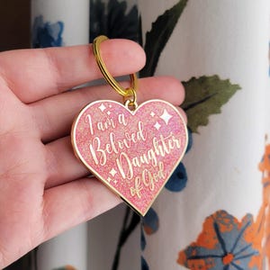 Puede incluir: Llavero de corazón rosa con purpurina dorada y las palabras "Soy una hija amada de Dios" en letras doradas.