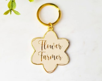 May include: Gold-coloured keychain with a white flower-shaped charm that says "Flower Farmer" in gold lettering.