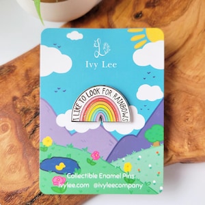 May include: A rainbow enamel pin with the text "I like to look for rainbows". The pin is on a blue and white card with a cartoon landscape and the text "Ivy Lee" and "Collectible Enamel Pins".