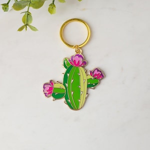 May include: A gold-tone keychain featuring a green cactus with pink and purple flowers. The cactus has three arms and is outlined in gold.