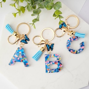 Könnte beinhalten: Drei bunte Schlüsselanhänger aus Harz mit einem blauen Schmetterlingsanhänger, einem blauen Quastenanhänger und einem goldenen Splitt-Ring. Die Buchstaben sind A, B und C. Die Buchstaben sind mit buntem Konfetti gefüllt.