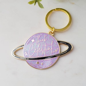 Op de afbeelding: Paarse glitter planeet sleutelhanger met een gouden ring en een zwarte ring rond de planeet. De planeet heeft de tekst "Think Celestial" erop.