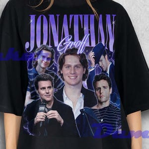 Puede incluir: Camiseta negra de gran tamaño con un collage de imágenes y el nombre "Jonathan Groff" en texto morado. El diseño incluye varias fotos del actor. La camiseta la lleva una persona.