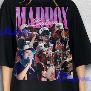 May include: Black oversized t-shirt featuring a graphic of a group of people with the text "MADDOX Batson" in pink. The graphic includes images of people wearing hats and jerseys. The shirt is worn by a person.