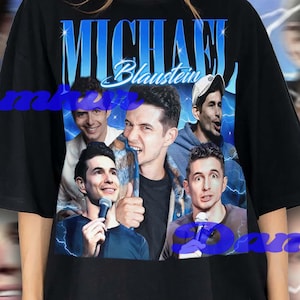 May include: Black t-shirt featuring a graphic of a group of people with the name "Michael Blaustein" in blue text. The background has a blue and white lightning design. The shirt is worn by a person.