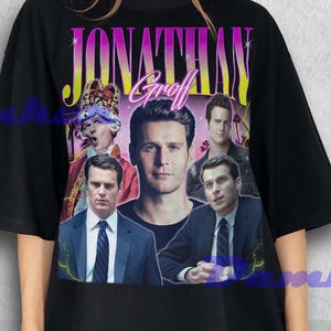 Puede incluir: Camiseta negra con un gráfico de Jonathan Groff y el nombre "JONATHAN Groff" en una fuente retro. El gráfico incluye múltiples imágenes del actor. La camiseta la lleva una persona.
