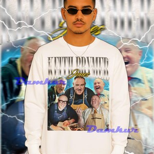 May include: White sweatshirt featuring a graphic of a group of men with the text "Keith Prymer Jones" in a vintage style. The design is set against a blurred background with lightning bolt accents.