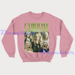 May include: Pale pink sweatshirt with a graphic of Caroline Forbes and the text "CAROLINE Forbes" in green. The graphic features multiple images of a blonde woman. The sweatshirt is a solid pale pink colour.
