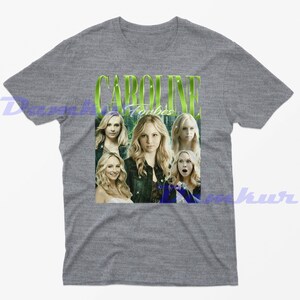 May include: Grey t-shirt with a graphic of Caroline Forbes and the text "CAROLINE Forbes" in green. The graphic includes multiple images of the character. The shirt is a short-sleeved crew neck.