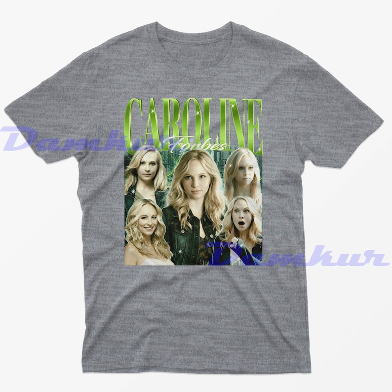 May include: Grey t-shirt with a graphic of Caroline Forbes and the text "CAROLINE Forbes" in green. The graphic includes multiple images of the character. The shirt is a short-sleeved crew neck.
