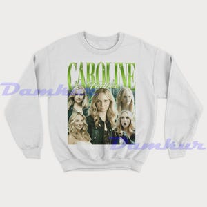 May include: White sweatshirt with a graphic of a woman with blonde hair, and the name "Caroline Forbes" in green text. The design includes multiple images of the same woman. A fan-made item.