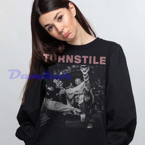 Puede incluir: Sudadera negra de manga larga con una imagen de concierto y la palabra "TURNSTILE" en una fuente texturizada de color rosa. La imagen muestra a una banda actuando en el escenario. La modelo lleva la sudadera.