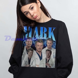 K&ouml;nnte beinhalten: Schwarzes Sweatshirt mit einem Aufdruck von Mark Sloan und dem Text "MARK Sloan" in Blau und Wei&szlig;. Das Design zeigt mehrere Bilder der Figur. Das Sweatshirt hat einen Rundhalsausschnitt.