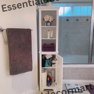 May include: White bathroom cabinet with three shelves and a door. The top shelf has a decorative flower arrangement and a bottle of perfume. The second shelf has a diffuser and two bottles of lotion. The third shelf has a purple towel. The bottom shelf has a variety of items, including a roll of toilet paper, a bottle of spray, and a box of tissues. The cabinet is open and the door is propped open.