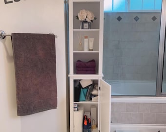 May include: White bathroom cabinet with three shelves and a door. The top shelf has a decorative flower arrangement and a bottle of perfume. The second shelf has a diffuser and two bottles of lotion. The third shelf has a purple towel. The bottom shelf has a variety of items, including a roll of toilet paper, a bottle of spray, and a box of tissues. The cabinet is open and the door is propped open.