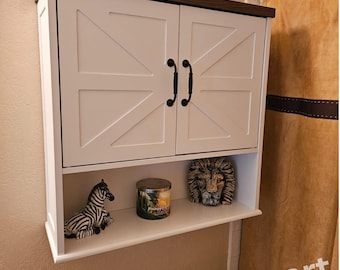 Könnte beinhalten: Weißer Badezimmer-Schrank aus Holz mit zwei Türen und einem Regal. Die Türen haben ein Design im Landhausstil mit schwarzen Metallgriffen. Der Schrank ist an einer Wand montiert und hat eine dekorative Holzplatte. Das Regal ist mit einer Zebra-Figur, einer Kerze und einer Löwen-Figur dekoriert.