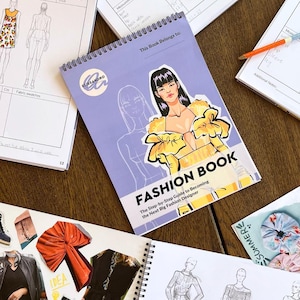 May include: A purple and white spiral bound book with the title "Fashion Book: The Step-by-Step Guide to Becoming the Next Big Fashion Designer". The cover features a colourful illustration of a woman wearing a yellow dress with large ruffles.