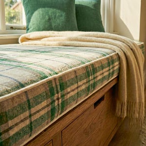 May include: A window seat with a plaid cushion in green, beige, and blue. Two green pillows rest against the window. A beige blanket with fringe is draped over the seat. The seat is made of wood.