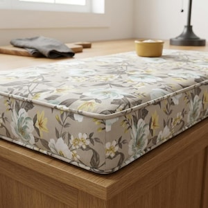 May include: A rectangular bench cushion featuring a light beige background with a floral pattern in shades of white, soft blue, and yellow. The cushion has piped edges and a structured, boxy shape, resting on a wooden surface.