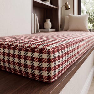 May include: A rectangular bench cushion featuring a woven houndstooth pattern in shades of deep red, black, and off-white. The cushion has a structured, box-edge design with clean, straight seams along the top and side borders, creating a firm profile.