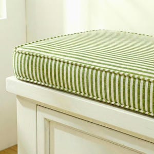 May include: A green and white striped bench cushion, with a white border, resting on a white wooden bench. The cushion's stripes are vertical. The design is simple and classic. The overall aesthetic is fresh and inviting. The cushion would fit a bench of approximately 76 cm.