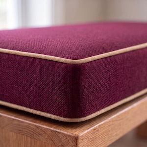 May include: A rectangular cushion upholstered in a deep burgundy, textured fabric with a prominent weave. The cushion features a light beige piping trim along the top and bottom edges, resting on a light-toned wooden base with a visible grain.