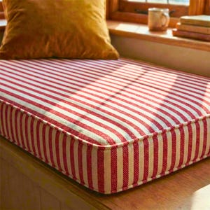 May include: A red and white striped cushion with a gold-coloured pillow in the background. The cushion is rectangular with a thick, padded border. The stripes are vertical and evenly spaced.
