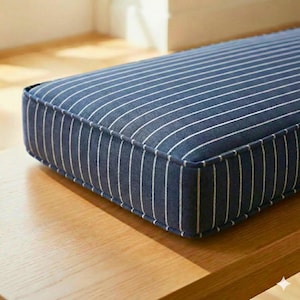 May include: A rectangular cushion with a navy blue and white striped pattern. The cushion has a thick, padded appearance with a contrasting border. The fabric has a textured look, and the cushion is placed on a wooden surface.