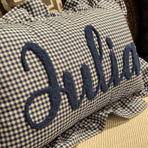 Könnte beinhalten: Ein rechteckiges Kissen mit blau-weißem Vichy-Muster. Das Wort "Julia" ist in einer strukturierten, marineblauen Schrift über die Mitte des Kissens gestickt. Das Kissen hat einen gerüschten Rand.