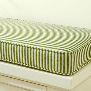May include: A rectangular cushion with a green and white striped design. The cushion has a white border and is placed on a white surface. The fabric has a textured appearance.