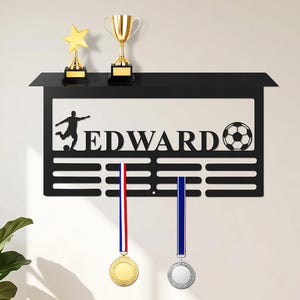 Op de afbeelding: Een zwart metalen wandrek met de naam EDWARD, een silhouet van een voetballer en een voetbalafbeelding. Het rek heeft een bovenplank met twee gouden trofeeën en meerdere horizontale sleuven voor het ophangen van medailles.