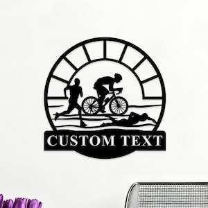 Puede incluir: Arte de pared de metal negro que representa una escena de triatlón. El diseño presenta un corredor, un ciclista y un nadador dentro de un marco circular. Las palabras "CUSTOM TEXT" se muestran en una pancarta rectangular.